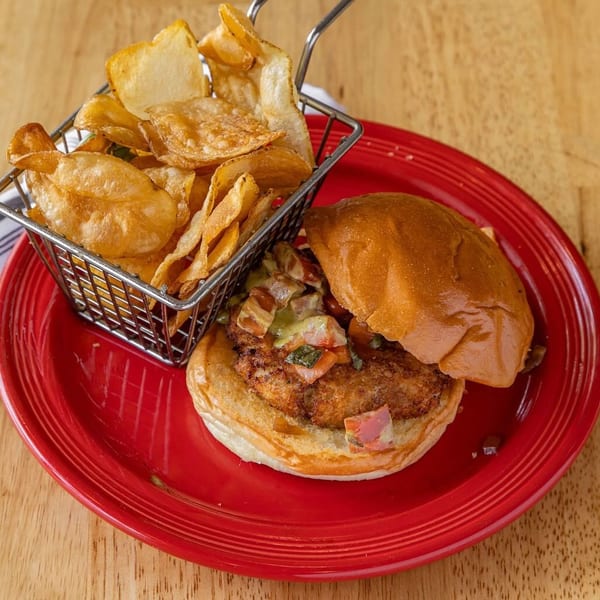 A chicken sandwich with salsa and a side of potato chips in a metal basket on a red plate.