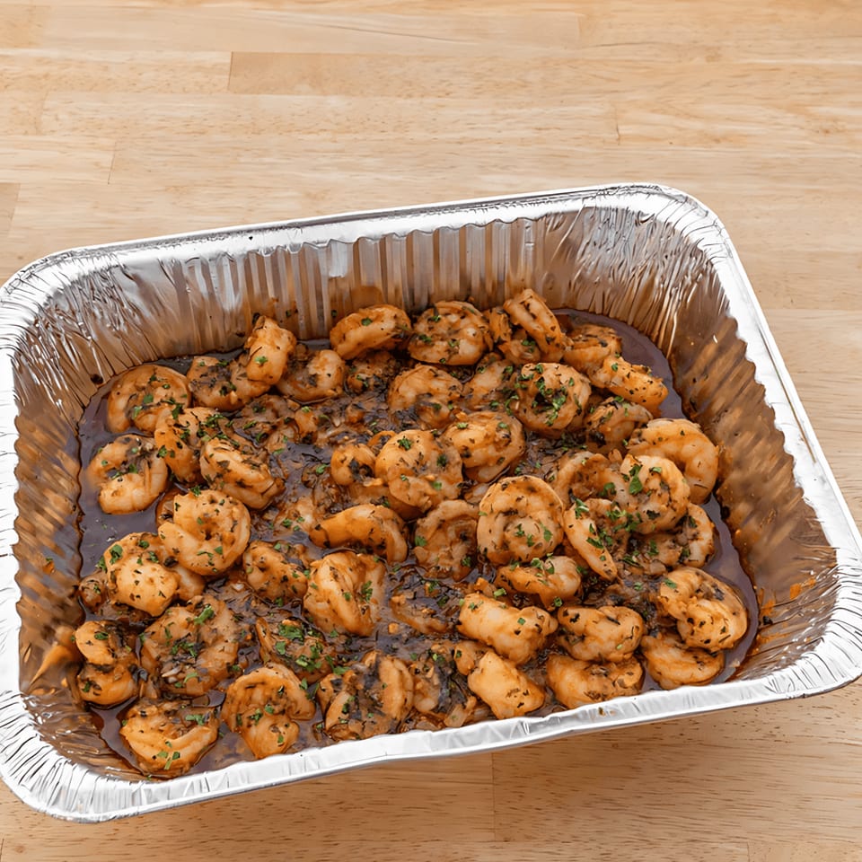 Shrimp scampi in a foil pan, garnished with parsley, on a light wooden table.