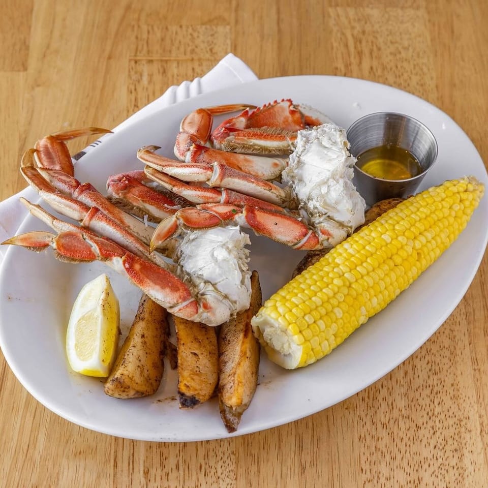 Snow crab legs, corn on the cob, potato wedges, lemon, and butter on a white plate.