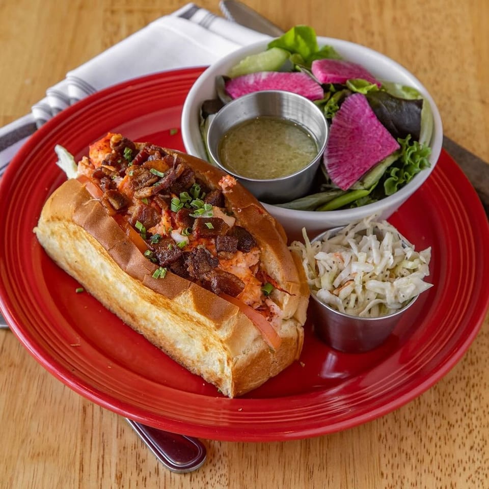 Lobster roll with bacon, coleslaw, and a side salad with watermelon radish on a red plate.