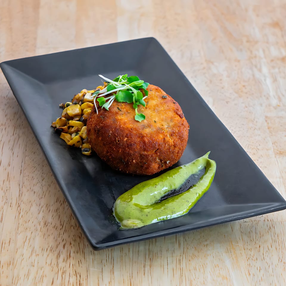 Crispy crab cake with roasted corn, microgreens, and green sauce on a black plate.
